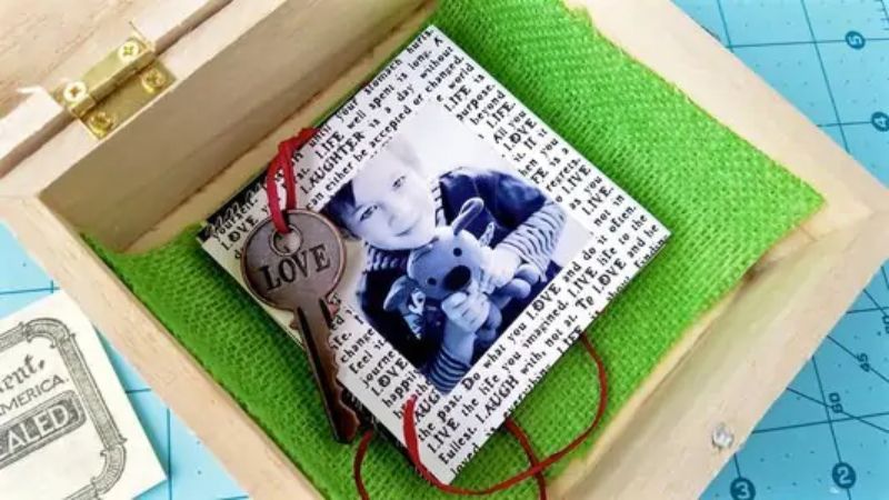 A wooden box with a green fabric lining contains a keychain with a round metal tag engraved with "LOVE" and a black and white photo of a child holding a stuffed toy. This perfect Mother’s Day gift has the photo resting on a piece of paper covered in text.