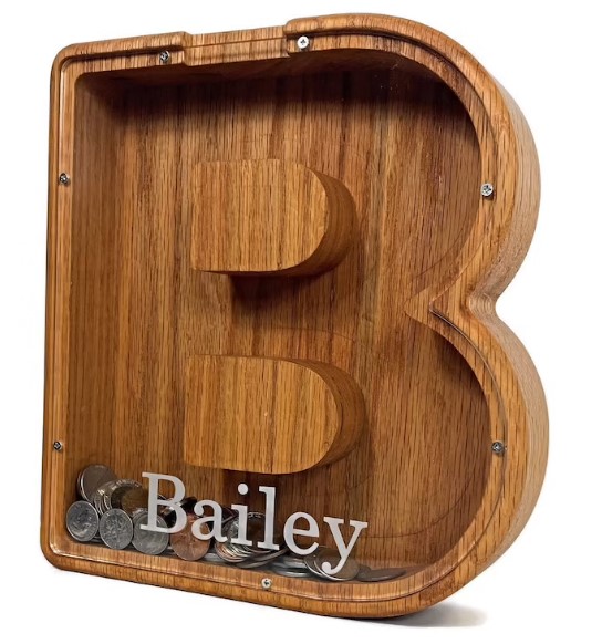 A wooden coin bank shaped like the letter "B" with a transparent front allowing coins inside to be visible. The name “Bailey” is written in white on the lower part of the clear front—an ideal gift idea for nieces and nephews of all ages.
