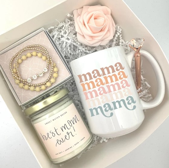 A gift box containing a white mug with "mama" written multiple times in pastel colors, a white candle labeled "Best Mom Ever!" in pink text, a set of beige and white beaded bracelets, a pen with a gem on top, and a pink rose—all on white shredded paper. Perfect Christmas gifts for new parents!