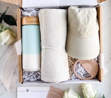 An open gift box contains a blue and white insulated bottle, a folded beige cloth, a beige baseball cap, and a light pink keychain with a sun design. Perfect for new parents, white roses and a calendar showing the number 9 are beside the box.