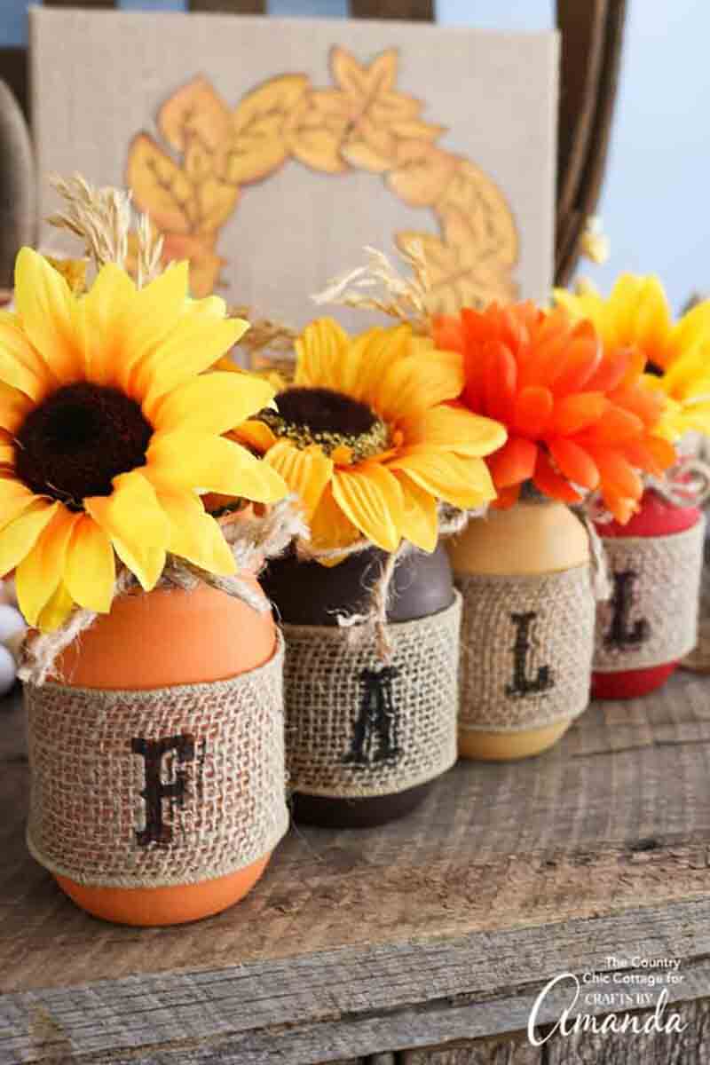orange, brown, yellow and red painted mason jars lined up with letters on burlap spelling out the word Fall