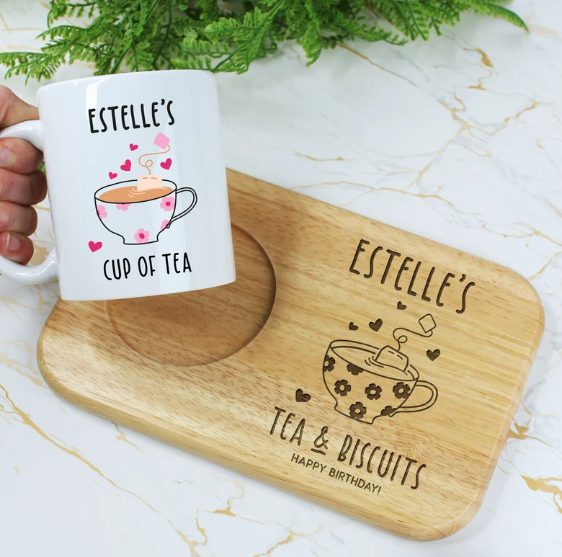 A hand holds a white mug labeled "Estelle's Cup of Tea" with a cute tea bag character and hearts. Below, a wooden serving tray with a teacup design and the text "Estelle's Tea & Biscuits" is shown. Both items, perfect gifts for seniors, rest on a white marble surface with green foliage in the background.