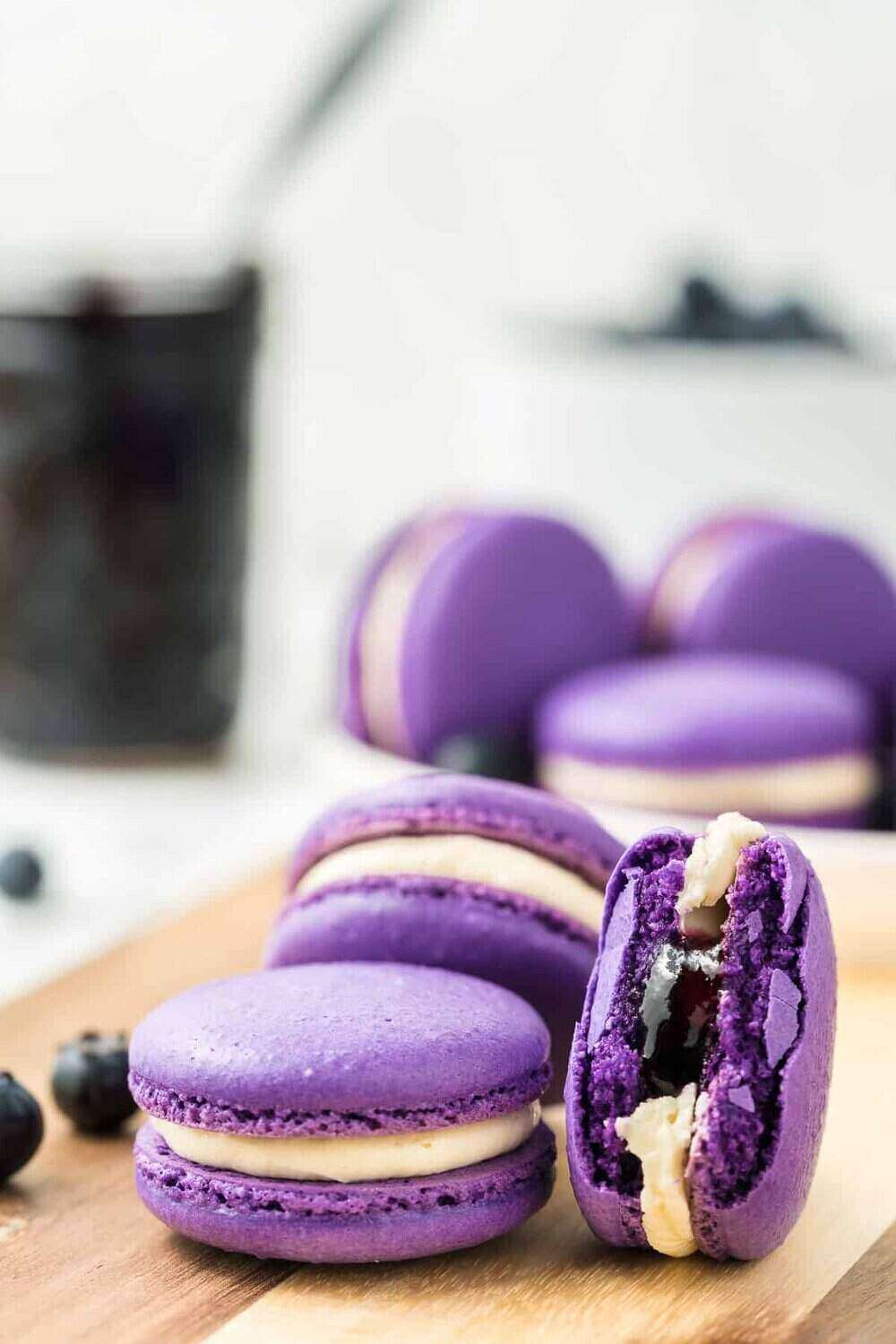 A photo of bright purple macarons with white frosting on a wooden cutting board. The front cookie has been pulled apart to show a blueberry filling. A cup of blueberries is unfocused in the back. 