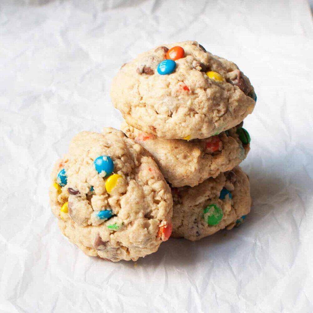 A table top with three stacked cookies sprinkled with colorful M&M candies. A fourth cookie leans on the side of the tower. 