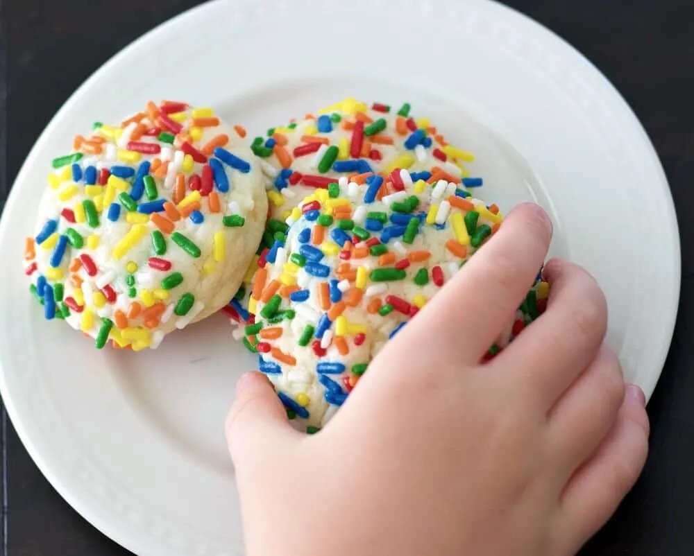 A white plate with colorful sprinkle covered cookies perfect for a summer bake sale. A child's hand is reaching to pick up one of the cookies. 