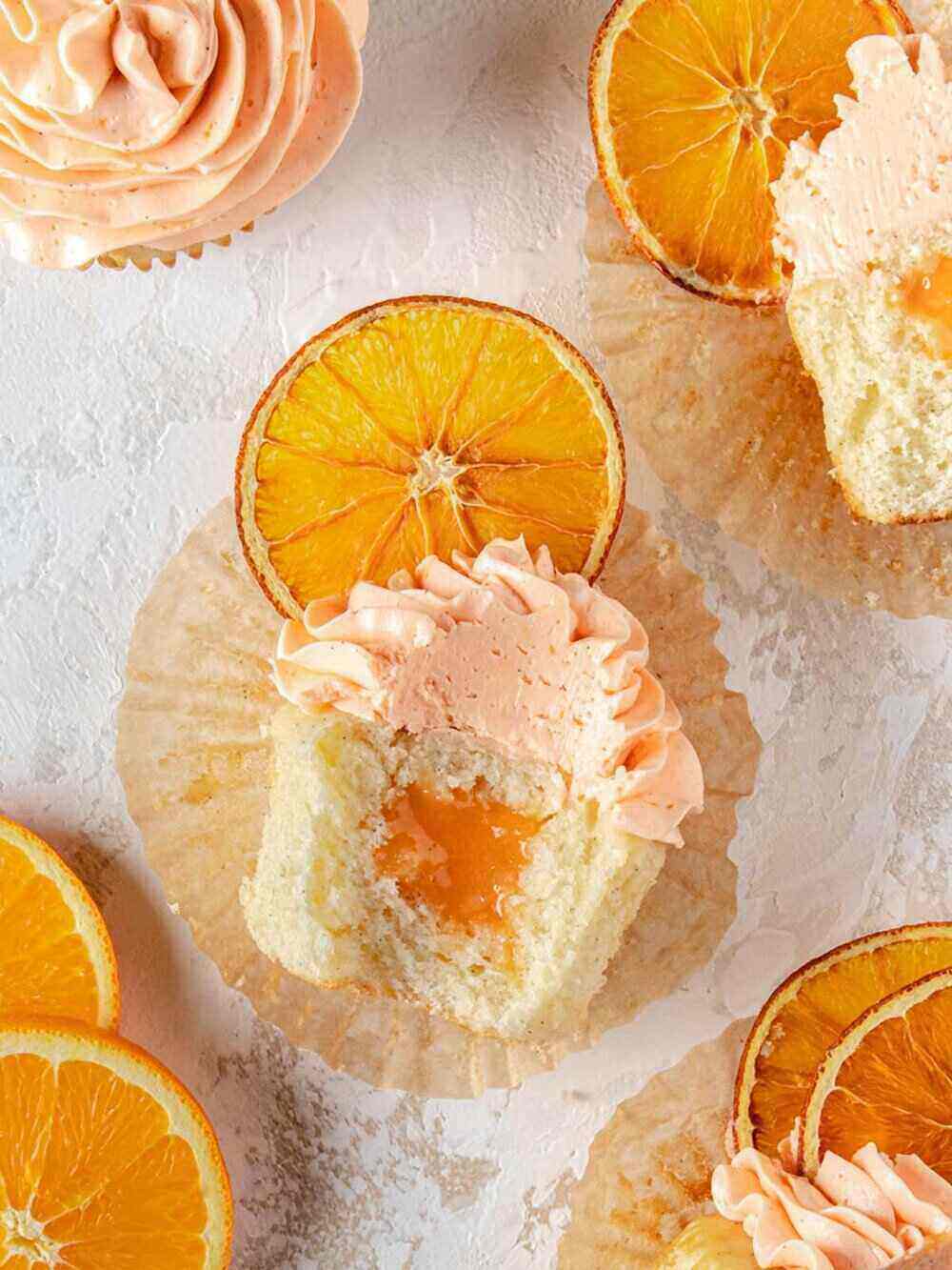 An unwrapped cupcake sits atop a cupcake liner. The cupcake is open to show an orange filling. Dried oranges decorate the photo along with other orange creamsicle cupcakes. 