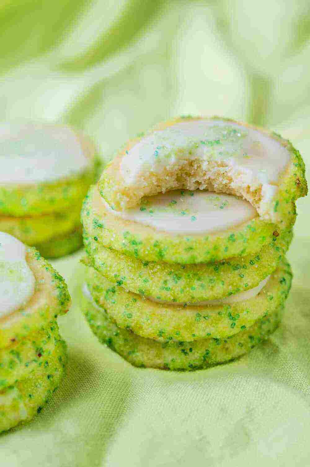 A photo of a stack of green key lime cookies with white frosting a green sprinkles sit on a light green tablecloth. 