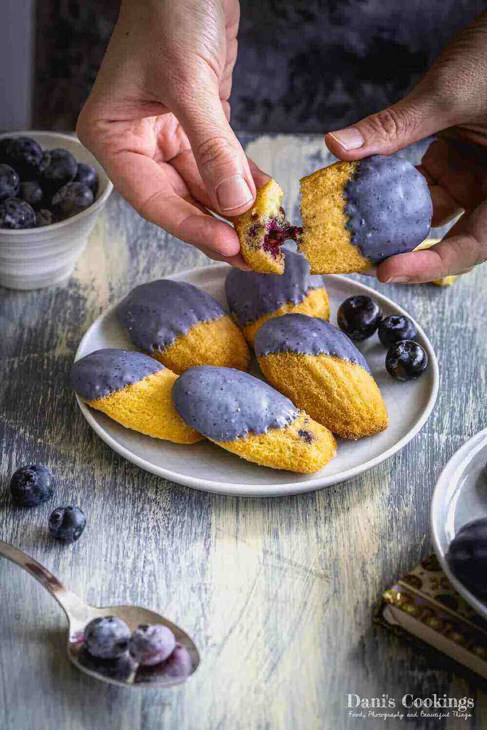 A photograph of a gray wooden table with a plate containing five Madeleine cookies dipped in purple white chocolate. Blueberries are scattered on the table and a person's hands are tearing the cookie into two pieces revealing blueberries. 