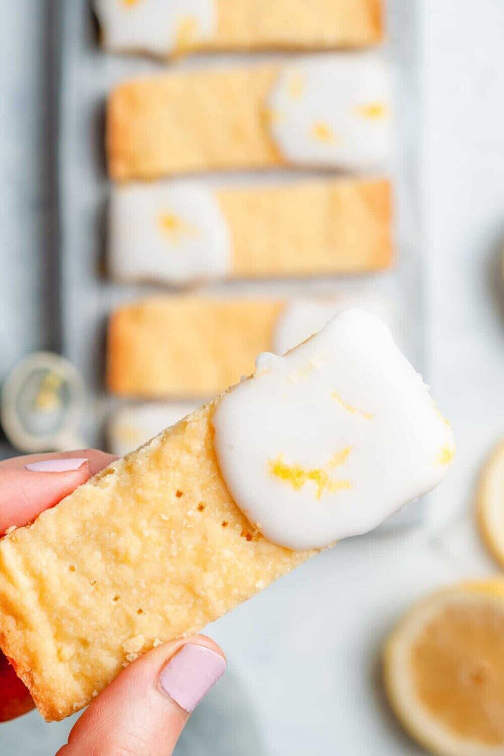 A photo of a white table cloth with iced lemon shortbread cookie bars. A hand holds one cookie bar towards the camera. 