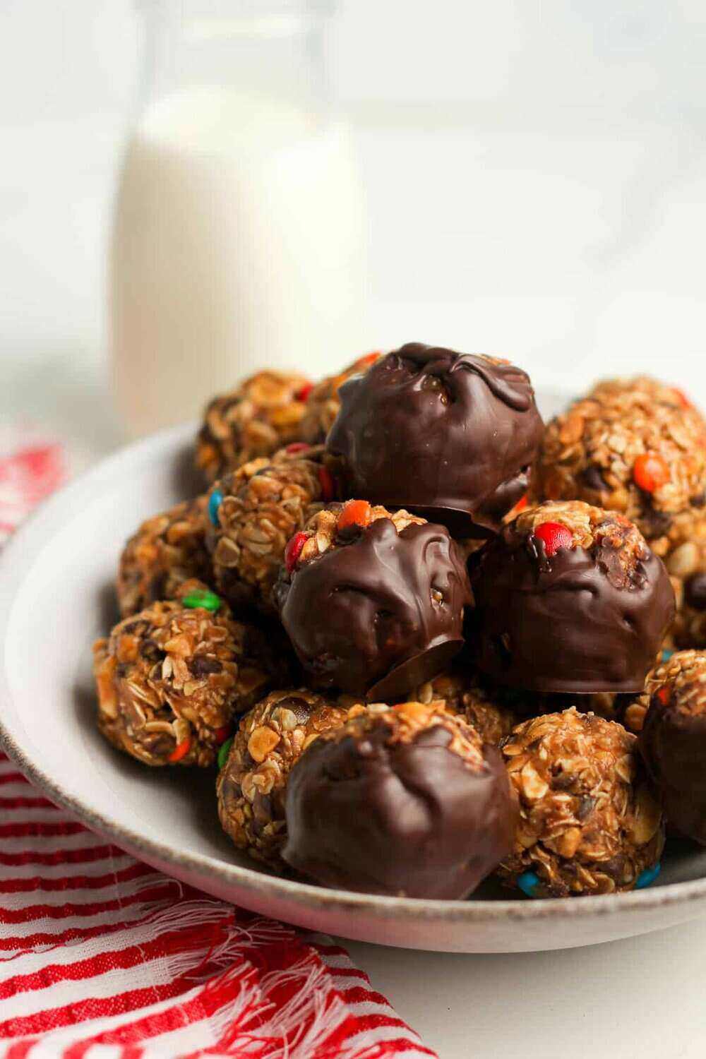 A plate filled with balls of no-bake cookie, dipped in chocolate. A red striped napkin is underneath the plate and a glass of milk stand behind it. These are delicious and make a simple snack for a summer bake sale idea. 