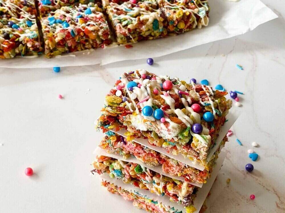 A stack of colorful rainbow Rice Krispie treats in front of a plate filled with them. Fruity Pebbles cereal lies sprinkled on the table.