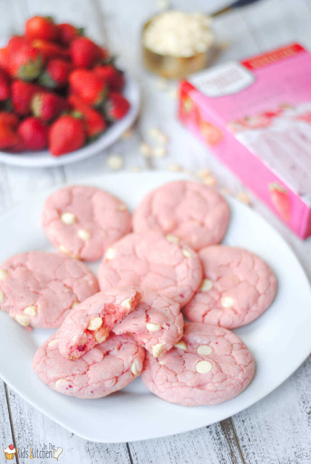 A photo of a plate of pink cookies with white chocolate chips sitting on a white plate. This summer bake sale idea also has a bowl of strawberries behind the plate. A strawberry cake mix box lies on the right side of the cookies with a full silver measuring cup behind it. 