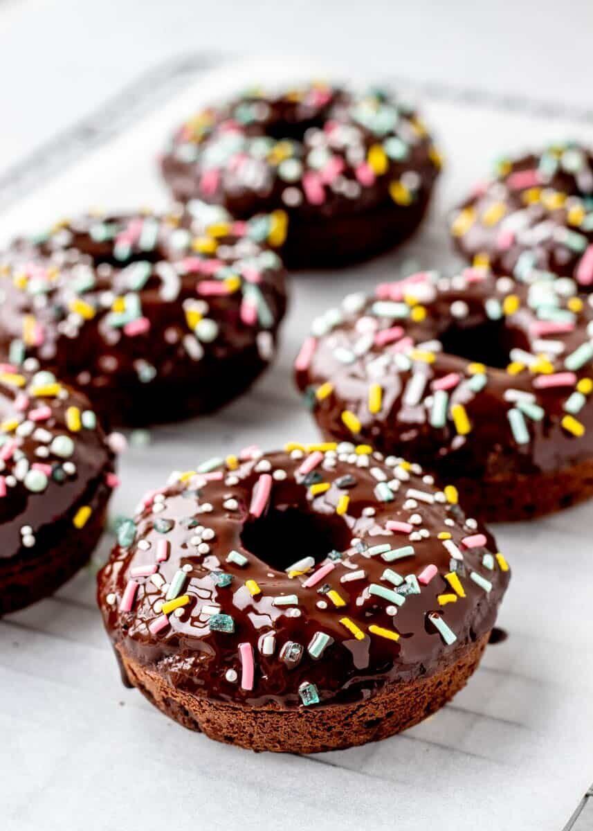 6 chocolate donuts with colorful sprinkles sit atop a cooling pan lined with parchment paper. 