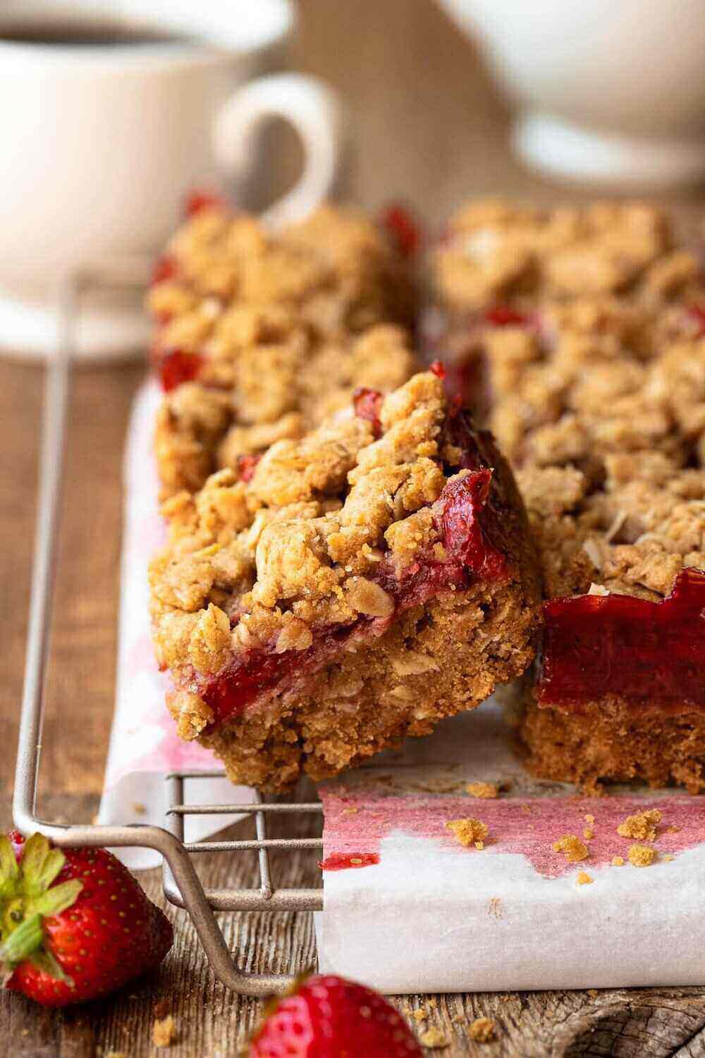 A sliced cookie bar with a fresh strawberry layer and crumble topping sits atop a cooling rack on a wooden table. Using fresh strawberries makes this an idea summer bake sale idea!