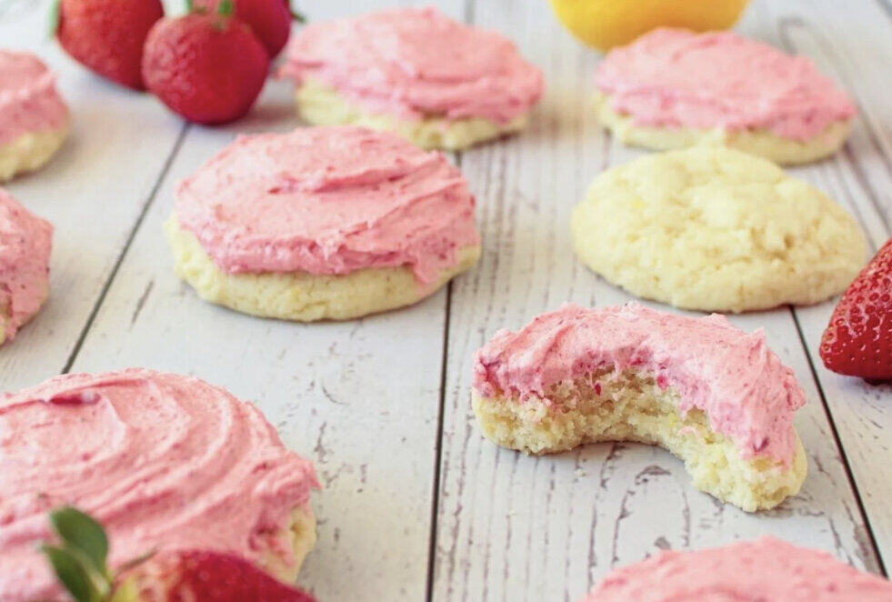 A wooden tabletop with pink frosted sugar cookies. There are strawberries sprinkled between the cookies. 