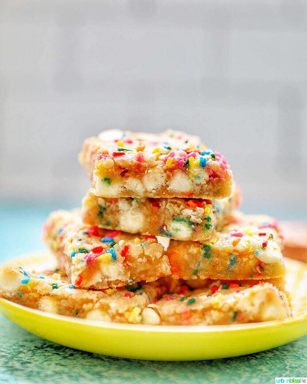 A photo of a stack of colorful sprinkle and white chocolate chip cookie bars on a yellow plate. The plate sits on a blue/green table.