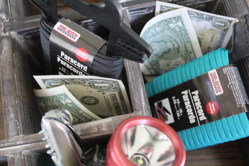 A brown box with multiple compartments holding emergency items of paracord, cash, a flashlight and a spork. 