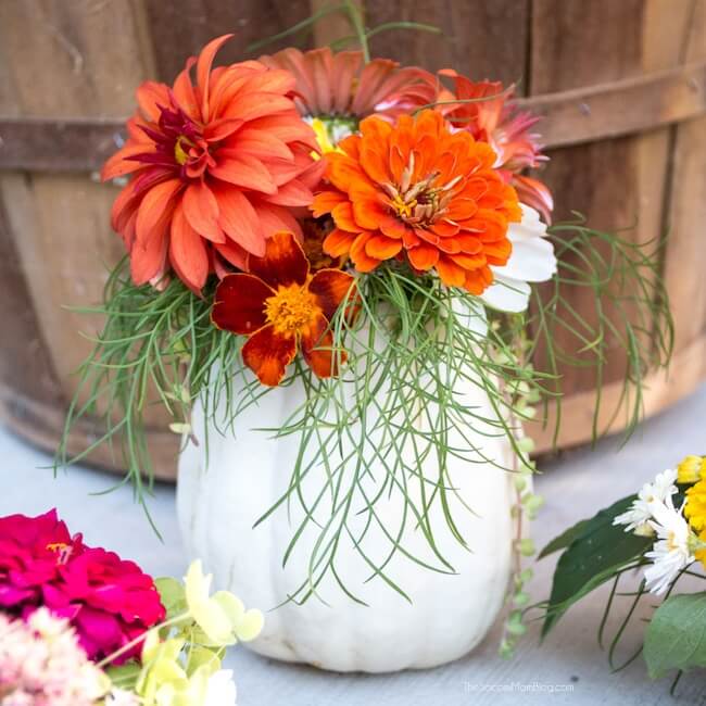 A brown barrel sits in the background. In front of it, a mini white pumpkin has beautiful green, orange and red flowers inside of it. 
