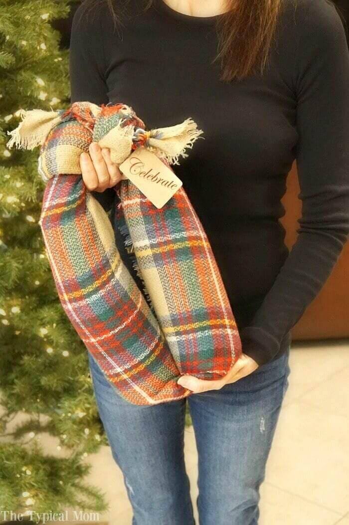 A photo of a woman standing beside a Christmas tree. She is holding a read plans parcel that has two wine bottles inside of it.