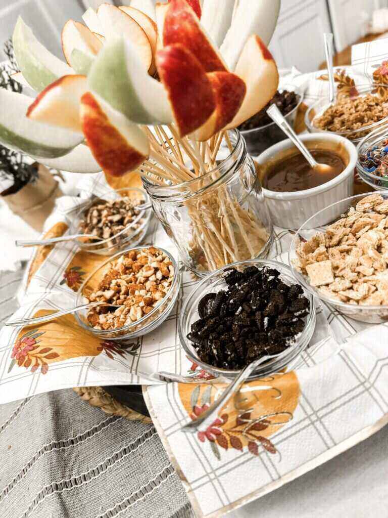 A photo of an apple dipping charcuterie board. A mason jar filled with apple slices on wooden dowels sits in the middle of a tray filled with glass bowls and toppings like caramel, nuts and Oreo cookies.