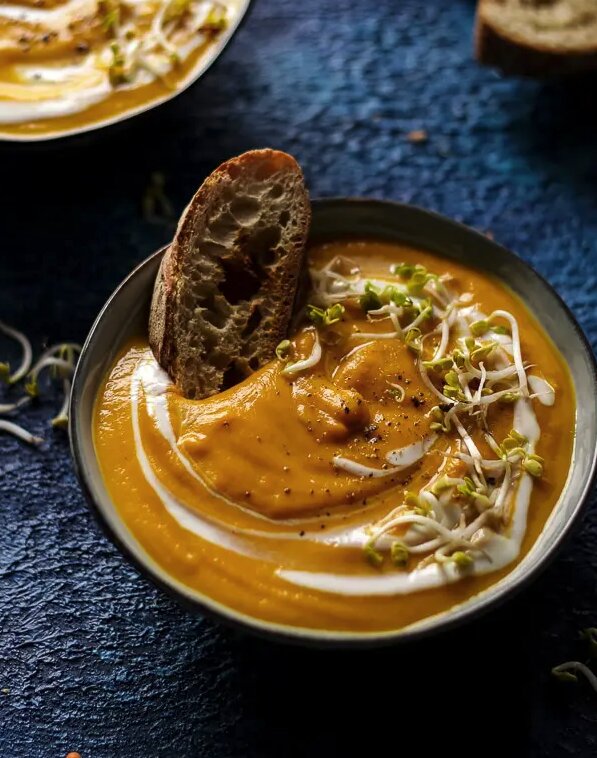 A blue countertop with a bowl full of thick creamy lentil soup with a piece of crusty bread stuck inside. 