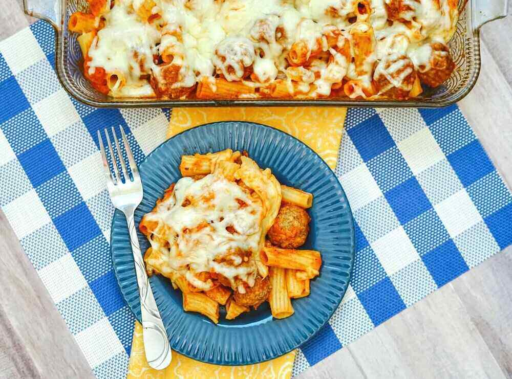 A photo of a meatball pasta bake in a casserole dish. It is on top of a blue checkered table cloth with a blue bowl and fork. 