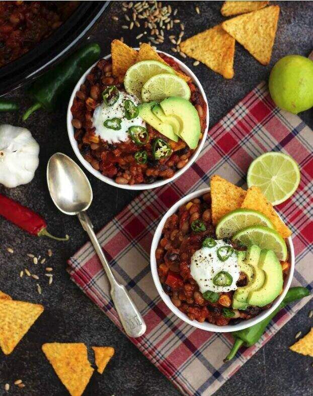 Two bowls of chili with lime wedges and tortilla chips sit on top of a table with red checkered napkins. 