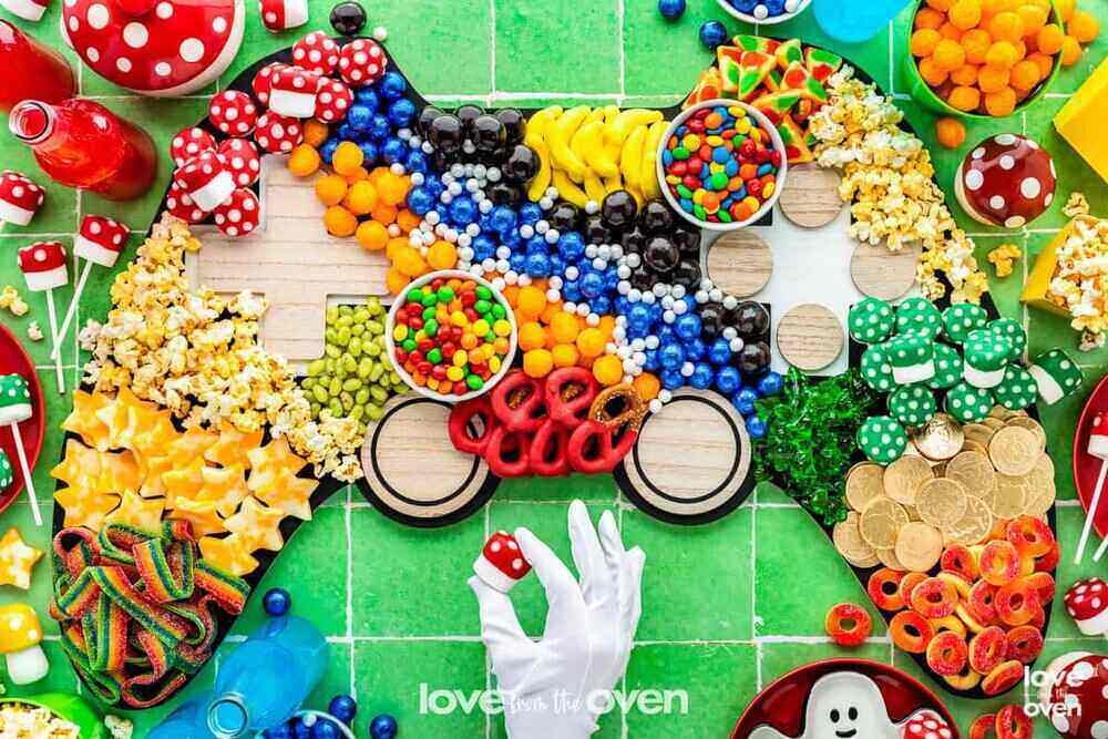 A colorful green tablecloth with candy and snacks in a Super Mario Bros Charcuterie. A hand with a white glove holds a small mushroom shaped marshmallow.