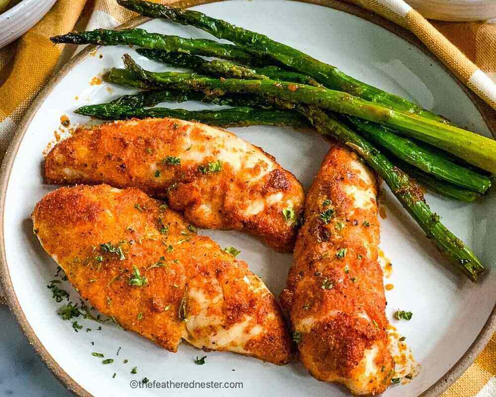 A white plate with three seasoned chicken tenderloins and cooked asparagus. 