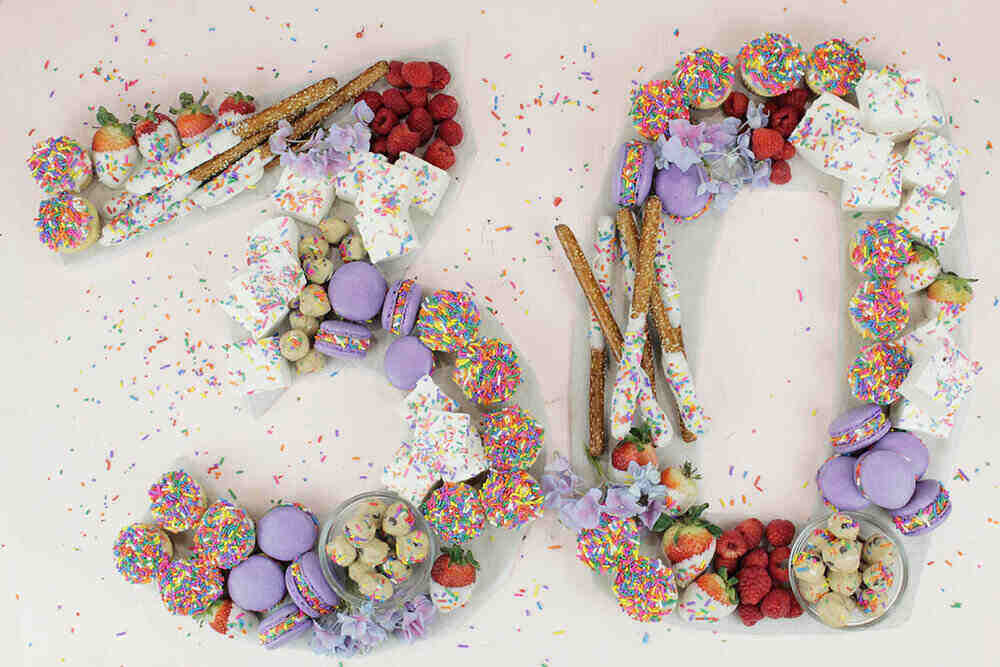 An array of colorful desserts, including macarons, strawberries, pretzels, raspberries, and assorted sweets, arranged to form the number "50" on a white background with scattered sprinkles—perfect for unique charcuterie boards at birthday parties.