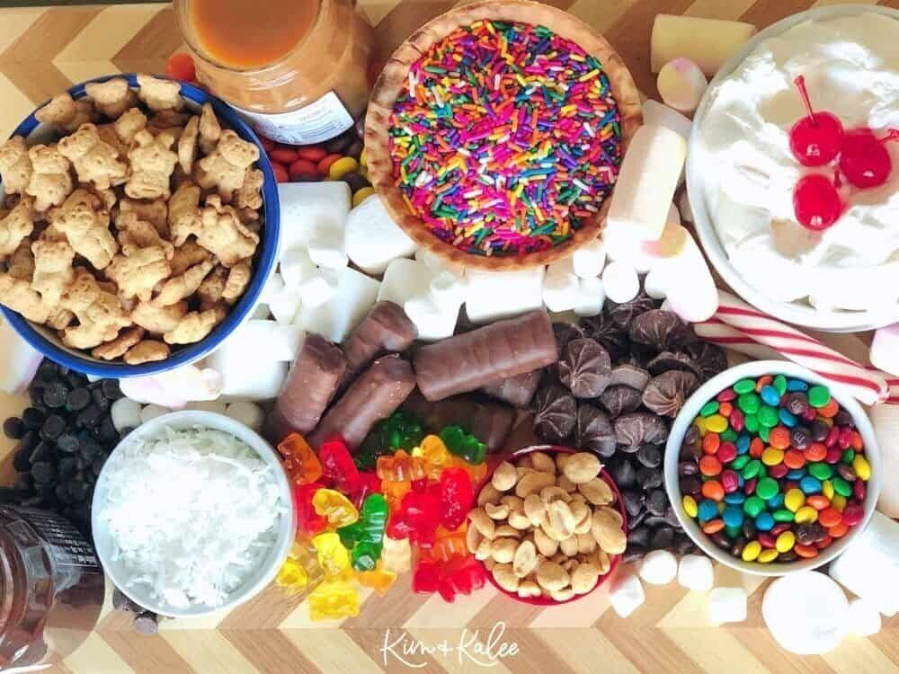 An overhead photo with a wooden cutting board filled with ice cream toppings including sprinkles, teddy grahams, m and ms and other candy.