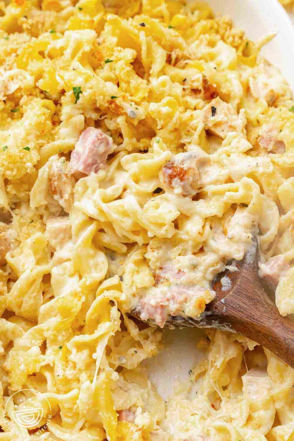 A dish of a creamy noodle casserole. A wooden spoon is scooping up the casserole. 