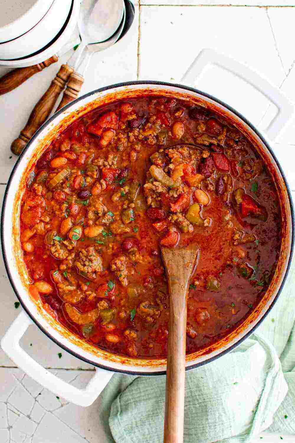 A large white pot with handles filled with tomatoes, beans and meat. A wooden spoon is in the middle. 