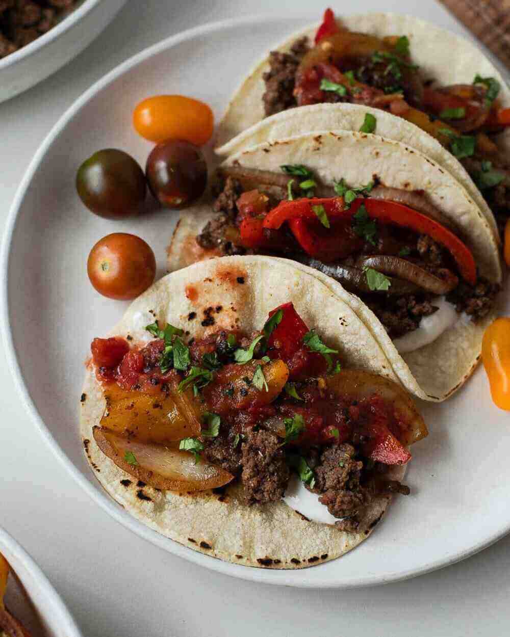 A white plate with three corn tortillas filled with ground beef, onions and peppers. 