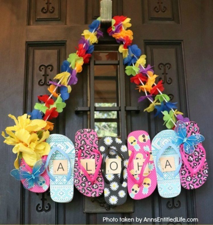 Colorful door decoration made of a lei of flowers hanging above flip-flops in different patterns and colors. Each flip-flop displays a letter, spelling out "ALOHA." Two blue butterflies and yellow flowers accent the decoration. Perfect for summer gift basket ideas!