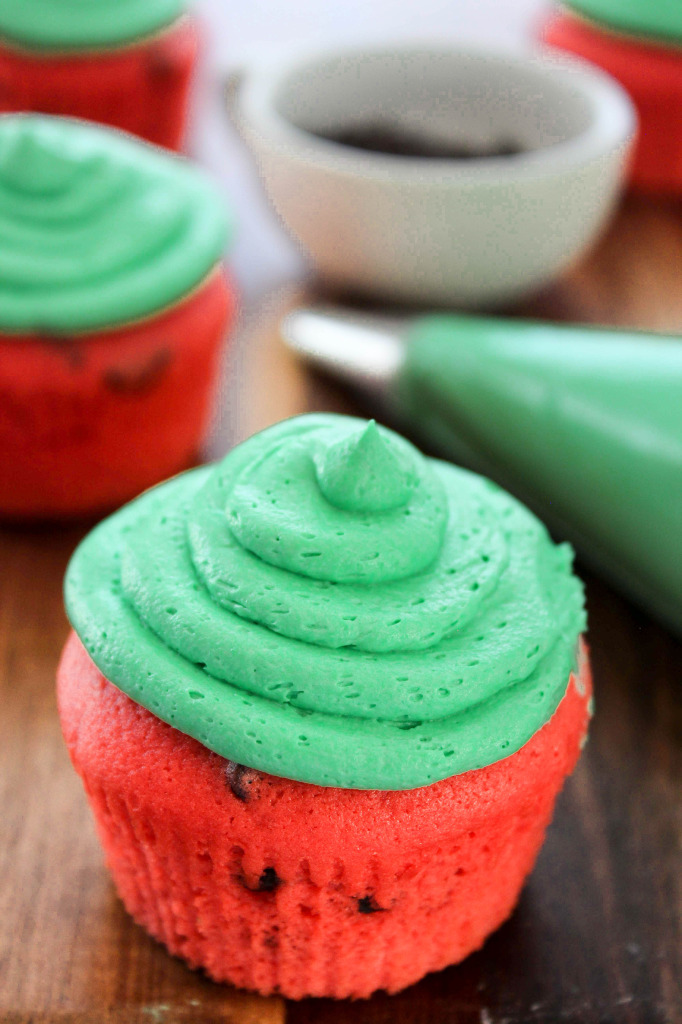 A close-up of a red cupcake topped with a swirl of green frosting sits on a wooden surface. Other similarly frosted cupcakes, a bowl, and a green frosting piping bag are in the background, perfect for adding to your summer gift basket ideas.