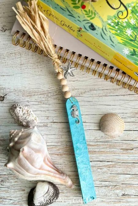 A turquoise bookmark with a small seahorse charm, wooden beads, and a raffia tassel lies on a wooden surface. Surrounding it are various seashells, and in the background, there is a closed spiral-bound notebook with a yellow-green cover. Perfect for summer gift basket ideas!