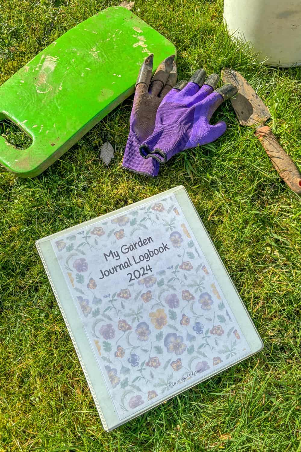 A "My Garden Journal Logbook 2021" binder with a floral cover lies on grass. Nearby, there is a green knee pad, a pair of purple gardening gloves, and a hand trowel. The scene suggests an outdoor gardening setting—perfect for summer gift basket ideas.