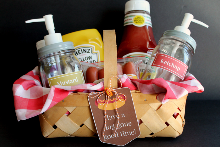 A picnic basket lined with red and white checkered cloth, containing mustard, ketchup, mason jars with pump dispensers labeled "Mustard" and "Ketchup," and a pack of hot dogs. A tag reads, "Have a dog gone good time!" It's one of the perfect summer gift basket ideas!