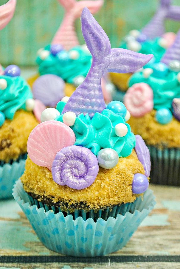 A cupcake decorated with a mermaid theme, featuring a purple mermaid tail made of fondant, teal and light purple frosting, and seashell-shaped confections. The base has a sandy texture made from crumbs, and the cupcake liner is blue with polka dots—perfect for summer gift basket ideas.