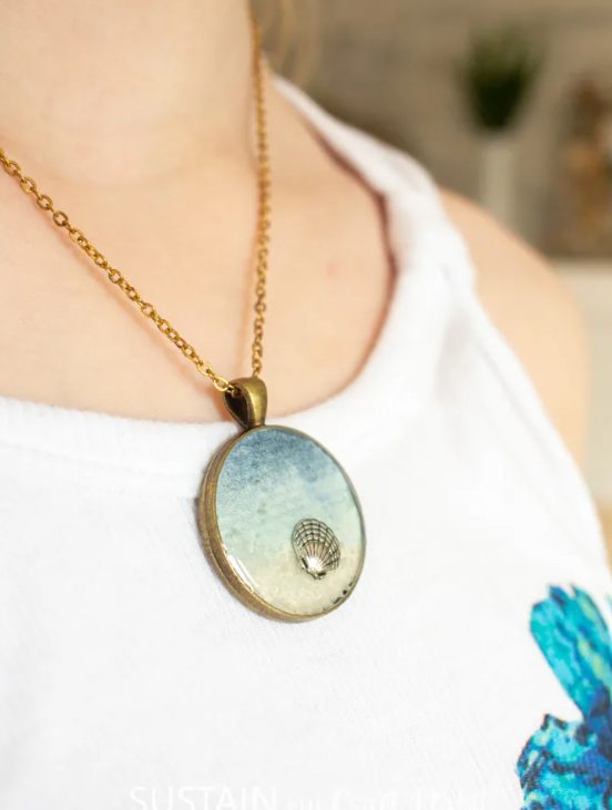 Close-up of a person wearing a round pendant necklace with an antique brass chain. The pendant features an artistic image of a hot air balloon floating in the sky—perfect for summer gift basket ideas. The person is also wearing a white tank top with a hint of blue design visible.
