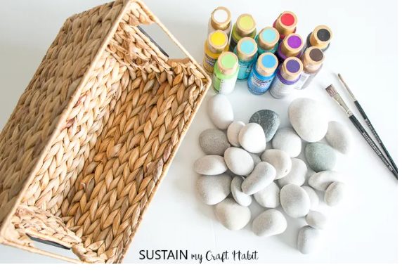 A woven basket is placed next to various art supplies on a white surface. Supplies include a collection of smooth grey stones, colorful bottles of paint, and a couple of paintbrushes. Perfect for summer gift basket ideas, the text "SUSTAIN my Craft HABIT" is at the bottom of the image.