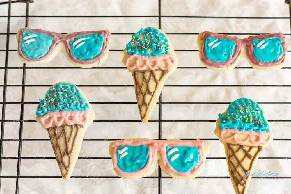 Colorful sugar cookies shaped like ice cream cones and sunglasses rest on a cooling rack. The ice cream cone cookies are decorated with blue and pink icing and sprinkles, while the sunglasses cookies are adorned with blue lenses and pink frames—perfect additions to summer gift baskets.