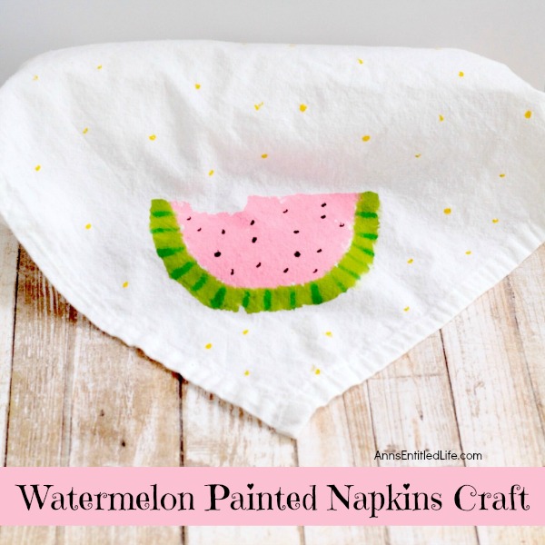 A white napkin is decorated with a hand-painted image of a watermelon slice, featuring a pink center, green rind, and black seeds. Small yellow dots are scattered around the napkin. The text "Watermelon Painted Napkins Craft" is displayed at the bottom—perfect for Summer Gift Basket Ideas.
