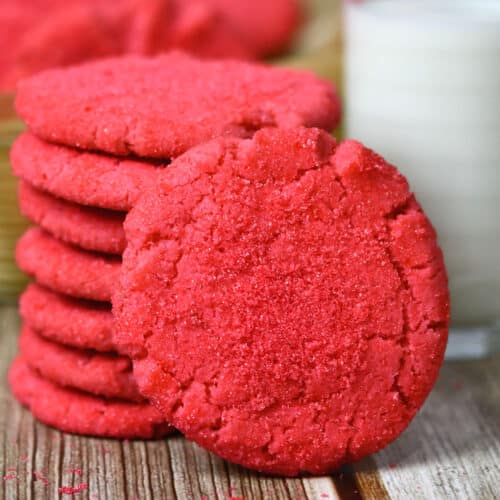 A stack of vibrant red cookies is arranged on a wooden surface, with one cookie leaning against the stack. In the background, a partially visible glass of milk adds a classic touch to the scene. These colorful baked goods boast a texture that appears soft and sugary, perfect for school bake sales.