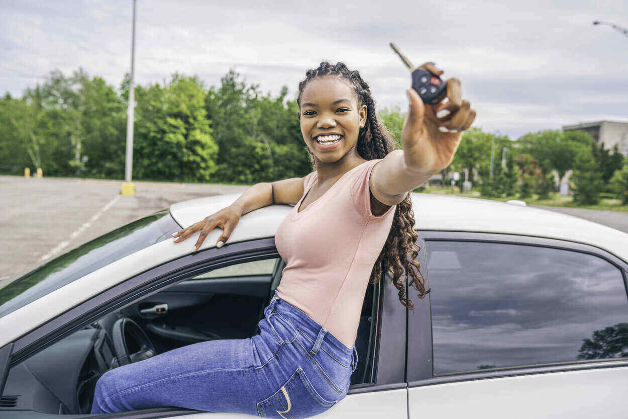 Teenage girl hanging out the driver side window smiling and holding a car key
