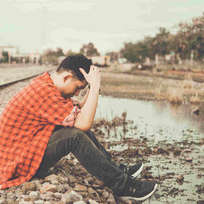 teenage boy sitting down next to a railroad track with his head in his hands like he is worried