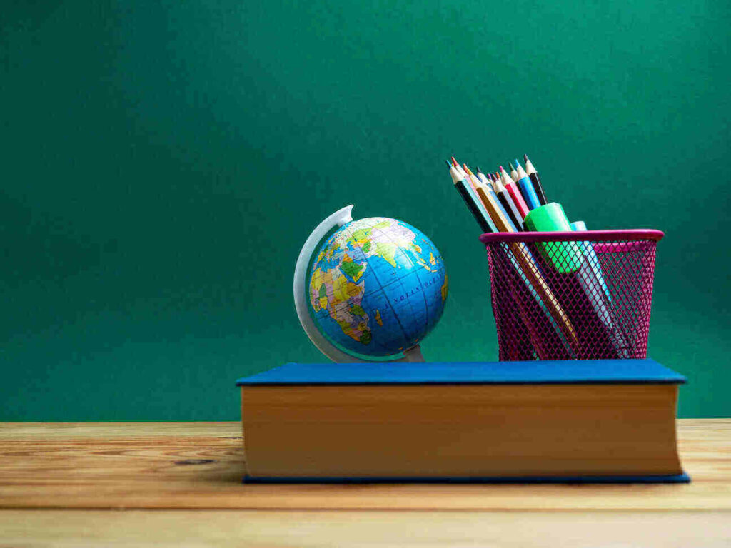 blue covered textbook sitting on a wooden desk in front of a small globe and pencil cup full of colored pencils