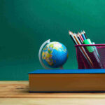 blue covered textbook sitting on a wooden desk in front of a small globe and pencil cup full of colored pencils